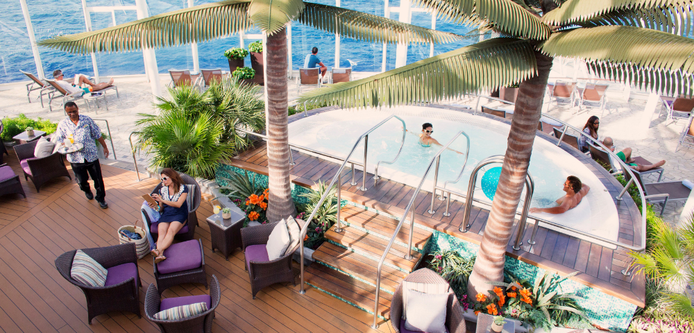 People relaxing on a cruise ship deck around a hot tub, surrounded by palm trees and lounge chairs with ocean in the background.