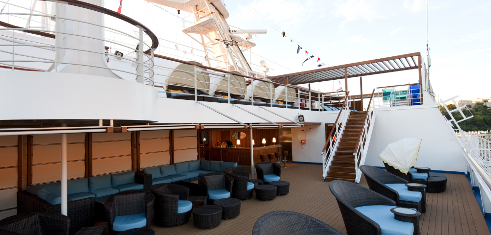 Outdoor lounge area on a ship with wicker chairs, blue cushions, stairs, and a bar under a covered deck.