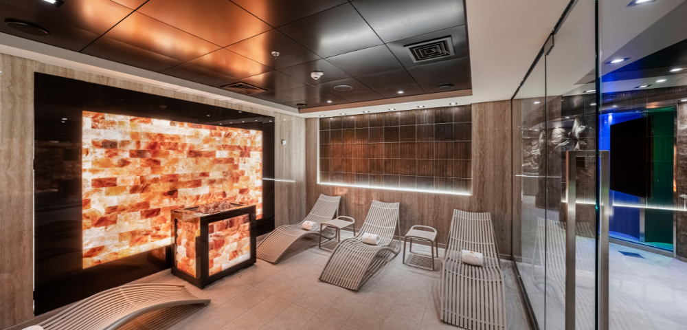 Modern spa relaxation room with illuminated salt wall, curved lounge chairs, towels, and glass door entrance.