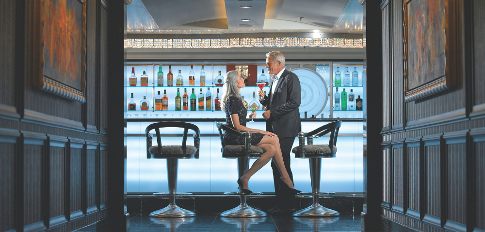 Mature couple enjoying drinks and conversation at a stylish, modern bar with illuminated shelves of liquor bottles.
