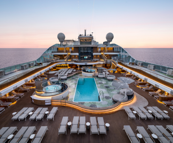 Luxury cruise ship deck at sunset with a central swimming pool, sun loungers, hot tubs, and ambient lighting overlooking the ocean.