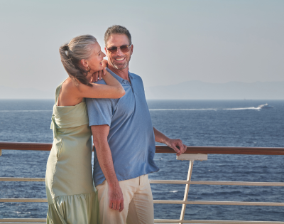 Couple smiling and embracing on a ship deck with a calm sea and distant mountains in the background.