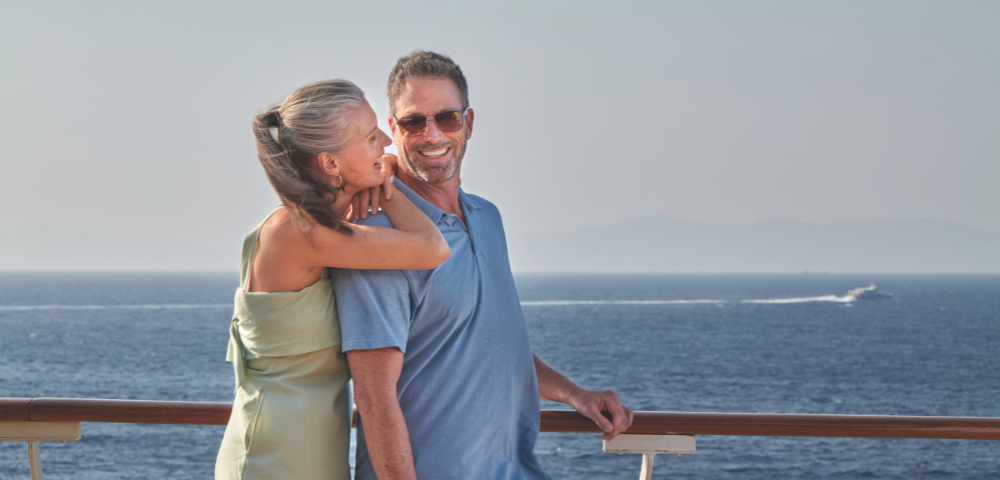 Couple smiling and embracing on a ship deck with a calm sea and distant mountains in the background.