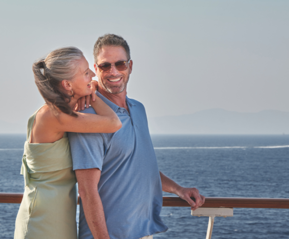 Couple smiling and embracing on a ship deck with a calm sea and distant mountains in the background.