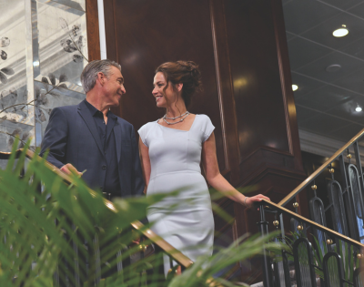 A man and woman in formal attire smiling and walking down an indoor staircase with plants and elegant decor.