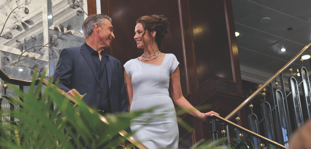 A man and woman in formal attire smiling and walking down an indoor staircase with plants and elegant decor.