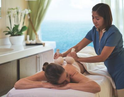 A woman receives a herbal ball massage on her back in a serene spa room overlooking the ocean.