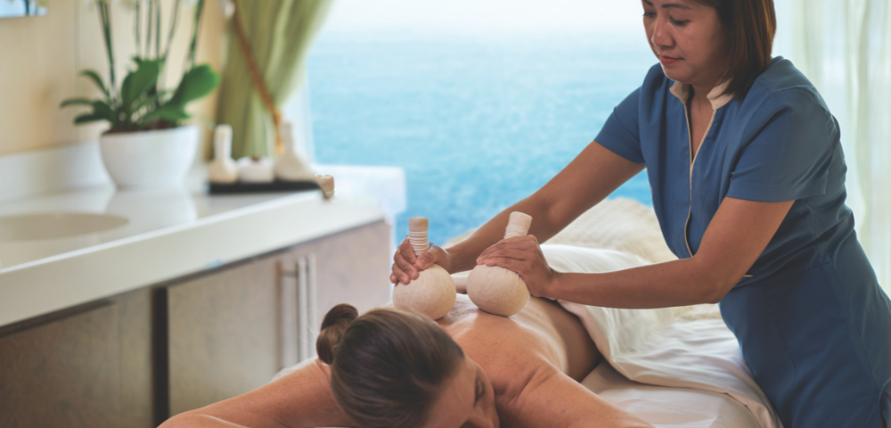 A woman receives a herbal ball massage on her back in a serene spa room overlooking the ocean.