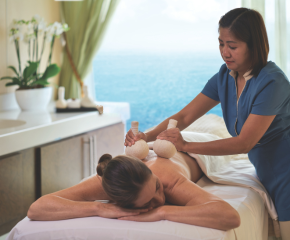 A woman receives a herbal ball massage on her back in a serene spa room overlooking the ocean.
