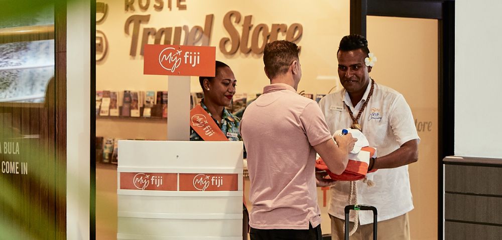 A man in a pink shirt interacts with two staff members at a travel store counter. They are smiling and handing him a package. The store has a welcoming atmosphere.