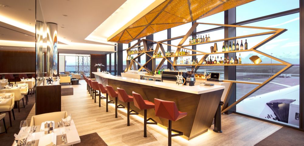 Modern airport lounge bar with a sleek, geometric design. Rows of red barstools line the white counter, while large windows reveal a runway view, creating a relaxing ambiance.