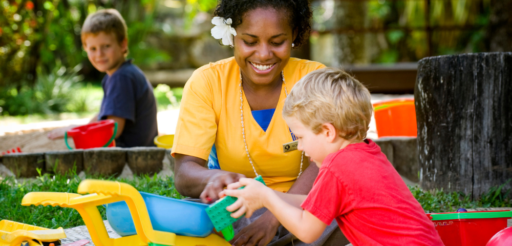 A woman in a yellow shirt and a child in a red shirt play with colorful toys outside. Another child sits in the background, with a playful and cheerful atmosphere.
