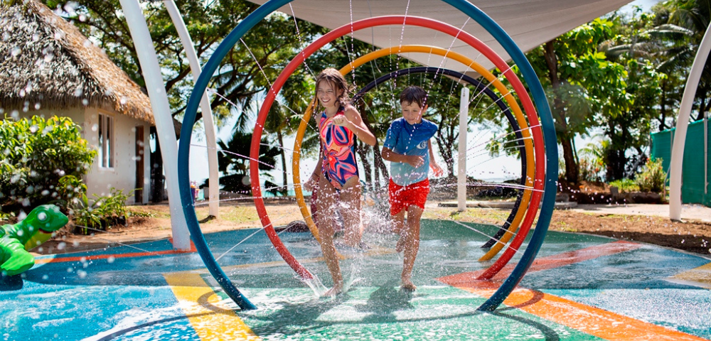 Children joyfully run through colorful sprinklers in a vibrant water park. Lush greenery and a thatched-roof hut create a playful, tropical setting.