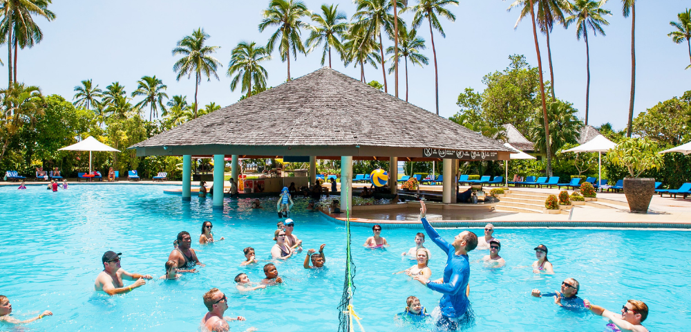 A lively pool scene at a resort, featuring a volleyball game with adults and kids. A thatched-roof bar is in the background, surrounded by palm trees.
