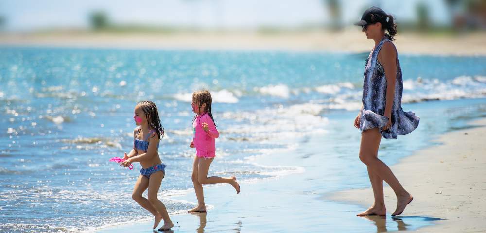 A woman and two children enjoy a sunny beach day. The girls, in swimsuits, run playfully by the shoreline, while the woman walks nearby, smiling.