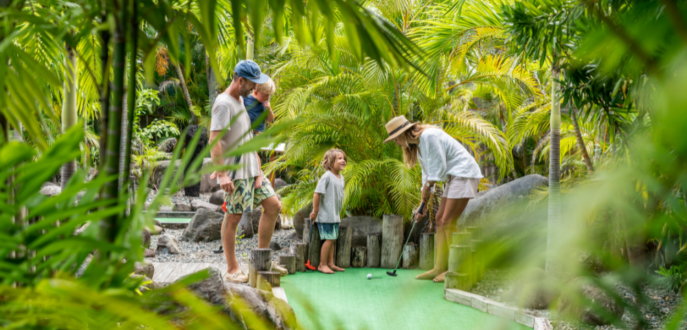 A family plays mini-golf surrounded by lush greenery. A woman in a sun hat lines up a shot as a child watches, creating a relaxed and joyful atmosphere.