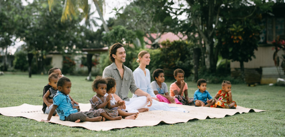 A group of adults and children sit on a mat in a lush garden, smiling and relaxed. The scene conveys warmth, community, and peaceful outdoor enjoyment.