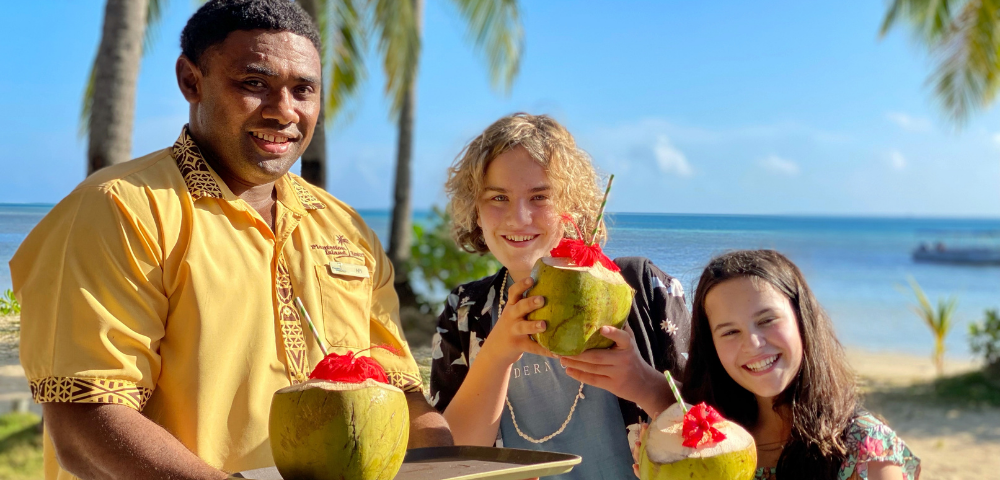 A cheerful man in a yellow shirt holds a tray with coconuts, standing with two smiling children by a beach. Palm trees and blue ocean are in the background.