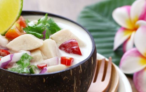 A coconut shell bowl of creamy Thai soup garnished with lime and cilantro, accompanied by vibrant plumeria flowers on a banana leaf, creating an exotic ambiance.
