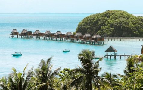 Overwater bungalows line a calm turquoise lagoon, with lush green islands and tropical foliage in the foreground and background under a clear blue sky.