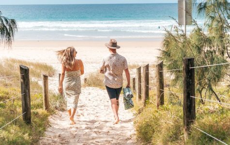 A couple walks barefoot on a sandy path toward a tranquil beach. The sky is clear, and green foliage lines the path, conveying a sense of serenity.
