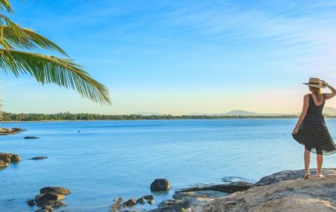 A woman in a polka dot dress and straw hat stands on a rock by the sea, gazing at the serene ocean and mountains under a blue sky.