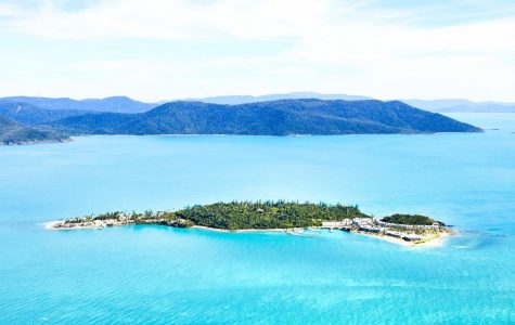 Aerial view of a lush, green island surrounded by vibrant turquoise waters and distant mountains under a bright blue sky.