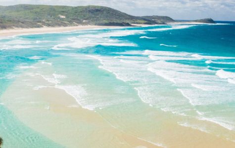 Aerial view of a pristine beach with turquoise waves, sandy shore, and lush green hills in the background, under a bright, cloudy sky.