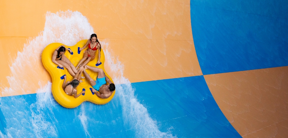 Four people ride together on a large yellow inflatable tube down a water slide with orange and blue sections.