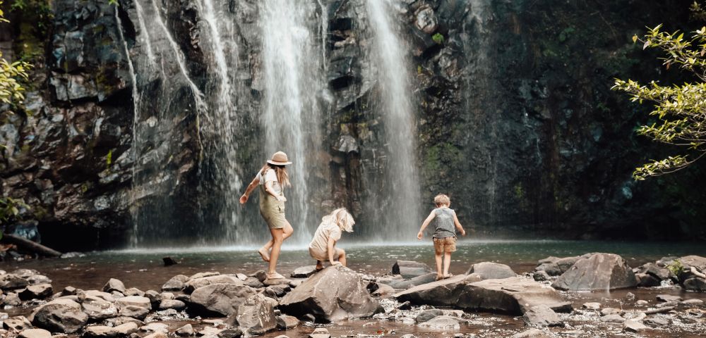 A woman and two children, explore rocks near a waterfall. The scene is calm and adventurous, set in a lush, natural environment.