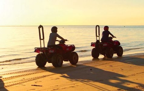 Silhouetted riders on ATVs cruising along a sandy beach at sunset, with calm water and a distant ship visible on the horizon.