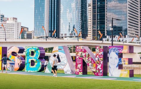 Colorful "BRISBANE" letters on grassy area, with people walking nearby. Modern skyscrapers and bridge in background, conveying a lively urban vibe.