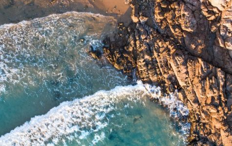 Overhead view of turquoise ocean waves crashing against a rugged, sunlit rocky coastline and a sandy beach.
