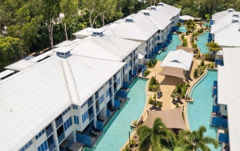 Aerial view of a resort with white-roofed buildings and turquoise swimming channels snaking between them, surrounded by lush green trees.