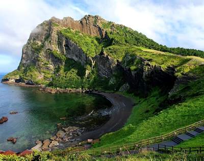 Rocky green hill overlooking dark ocean water with cloudy blue sky - Jeju Island South Korea