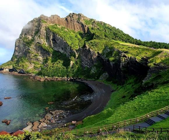 Rocky green hill overlooking dark ocean water with cloudy blue sky - Jeju Island South Korea