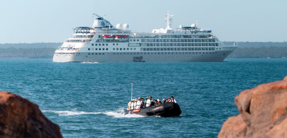 Small boat with passengers approaches rocky shore, large white cruise ship visible in calm blue sea in the background.
