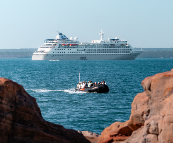 Small boat with passengers approaches rocky shore, large white cruise ship visible in calm blue sea in the background.