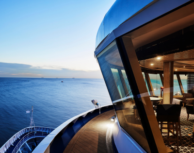 View from a ship’s deck overlooking calm ocean waters at sunset, with a modern indoor lounge area visible through large windows.