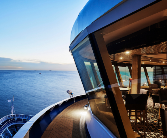 View from a ship’s deck overlooking calm ocean waters at sunset, with a modern indoor lounge area visible through large windows.