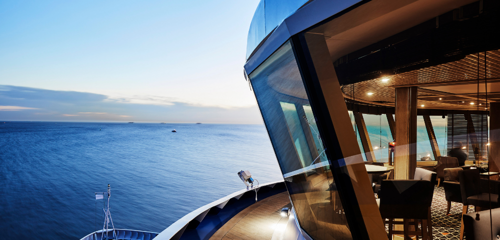 View from a ship’s deck overlooking calm ocean waters at sunset, with a modern indoor lounge area visible through large windows.