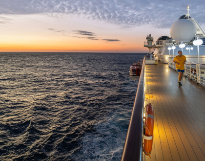 Person jogging on the deck of a cruise ship at sunset over a calm ocean with partly cloudy skies.