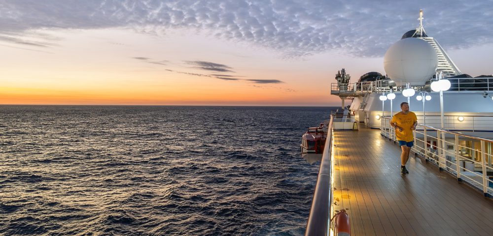 Person jogging on the deck of a cruise ship at sunset over a calm ocean with partly cloudy skies.