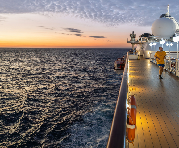 Person jogging on the deck of a cruise ship at sunset over a calm ocean with partly cloudy skies.