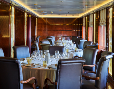 Elegant dining room on a cruise ship with round tables, leather chairs, and polished wooden walls and windows.