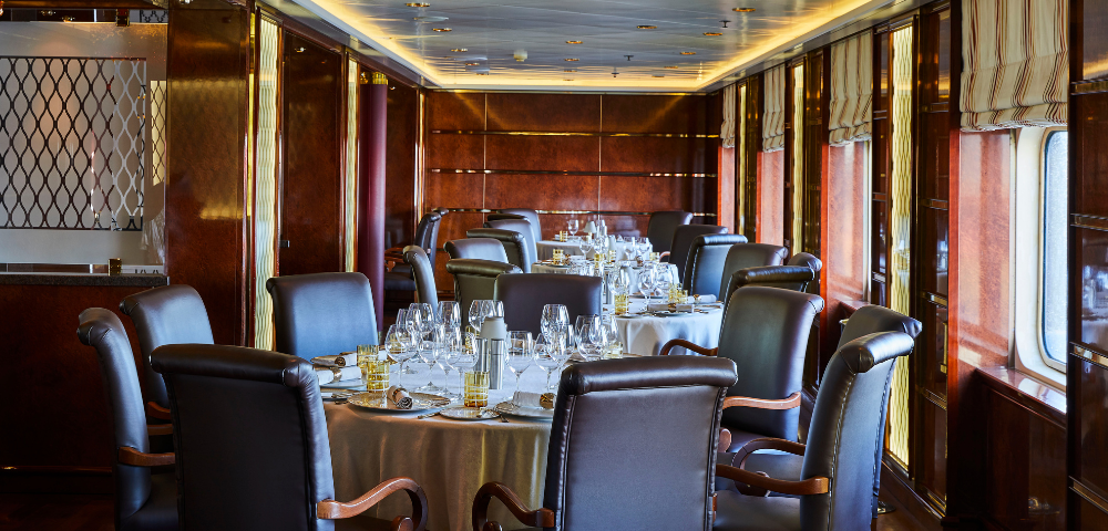 Elegant dining room on a cruise ship with round tables, leather chairs, and polished wooden walls and windows.