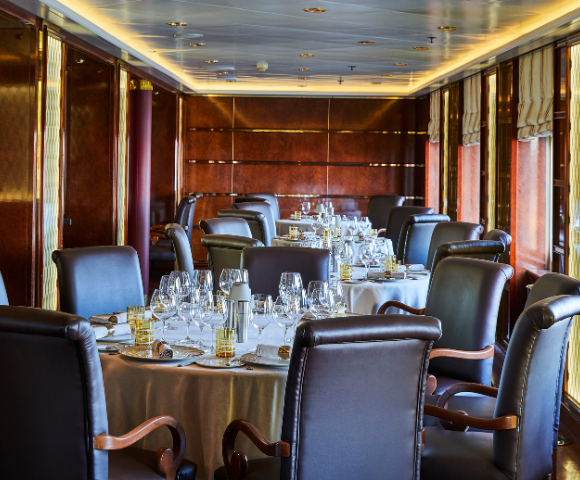 Elegant dining room on a cruise ship with round tables, leather chairs, and polished wooden walls and windows.