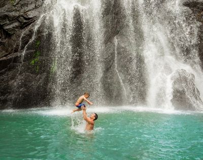father throwing his son up in the air while playing in the water in front of a waterfall