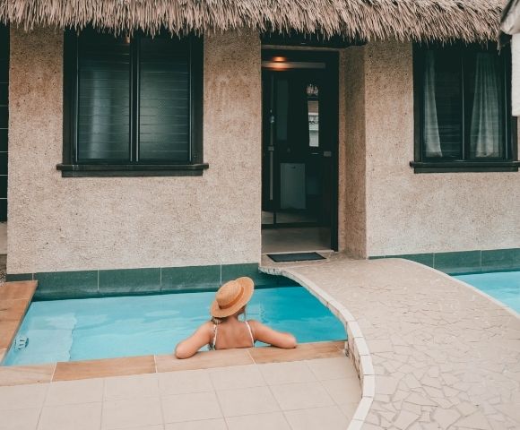 woman in hat enjoying private pool outside bure with small bridge and thatched roof