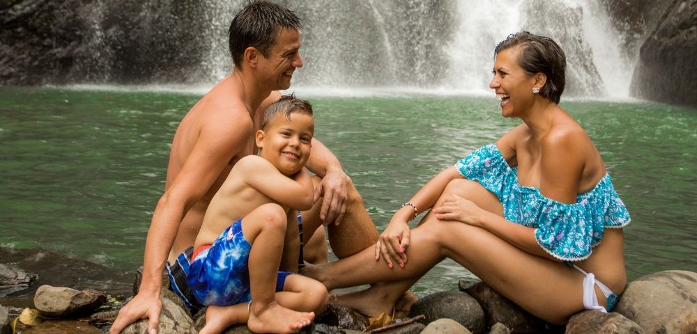 mother father and son sitting on the waters edge with a waterfall in the background